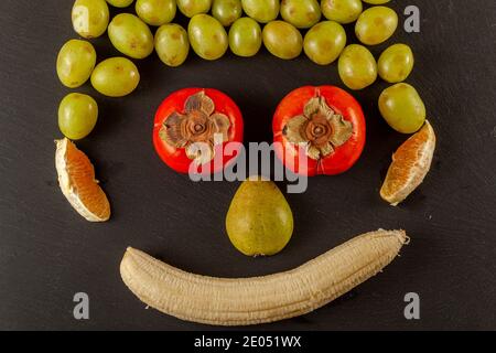 Pear, grapes, persimmon, oranges and peeled banana are placed on a black background to resemble human face. Flat lay image of an exotic fruit medley w Stock Photo