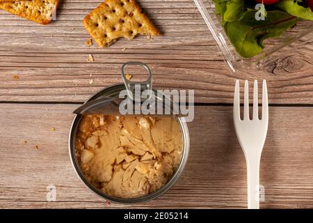 Flat lay image of quick and snack photo featuring a easy open can of tuna with tuna chunks in oil on wooden table or bench, clear plastic container wi Stock Photo