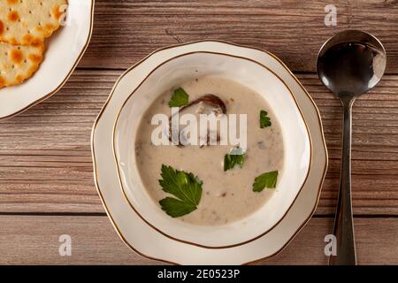 Flat lay image of Creamy mushroom soup decorated with parsley leaves in an artisan porcelain cup on a flat plate on a wooden table served with cracker Stock Photo