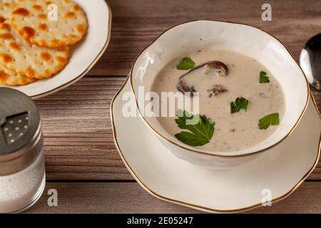 Side view of Creamy mushroom soup decorated with parsley leaves is in an artisan porcelain cup on a flat plate on a wooden table served with crackers, Stock Photo