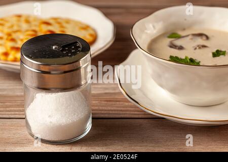A metal top glass salt shaker with cup of creamy mushroom soup and salty crackers in the background. An image for salt consumption heart and hypertens Stock Photo
