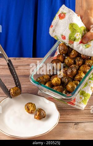 Close up image of a thick glass bowl of freshly cooked and seasoned Brussel sprouts being hold by a caucasian woman with kitchen cloth and served into Stock Photo