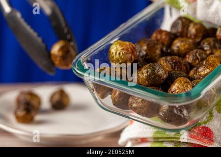 Close up image of a thick glass bowl of cooked and seasoned Brussel sprouts being held with kitchen cloth and served into porcelain plates using a ton Stock Photo