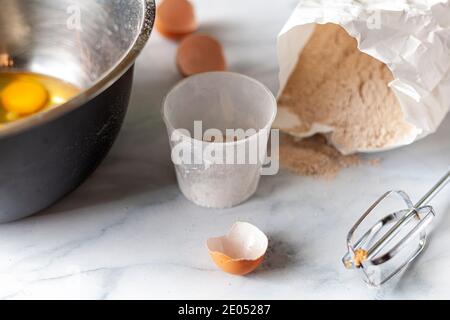 Pastry being prepared on marble kitchen countertop with a paper bag of wholewheat flour, broken egg shells, measuring cup and mixer attachment seen. R Stock Photo