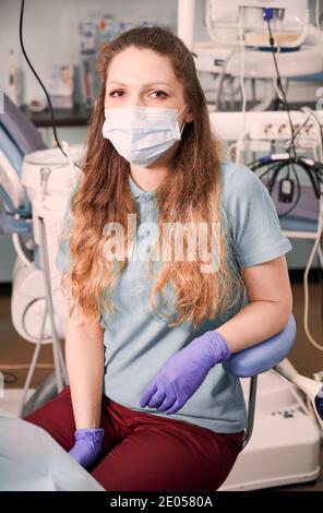 Portrait of charming female dentist in medical mask with long red hair posing in dental office with dental chair and special equipment. Concept of dentistry, healthcare and dental staff. Stock Photo