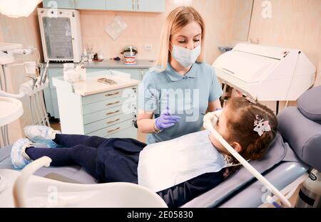 Young woman dentist in medical face mask holding dental instruments while little girl lying in dental chair with inhalation sedation. Concept of sedation dentistry. Stock Photo