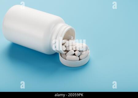 Vitamins and minerals in white capsules fell out of a white jar on a blue background. The concept of immune protection, antiviral prevention. Food add Stock Photo