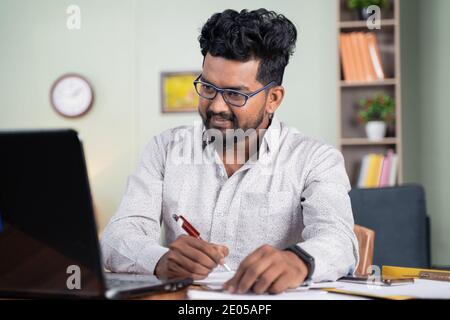 Young man writing down notes by looking laptop - concept of employee or student online training at home. Stock Photo