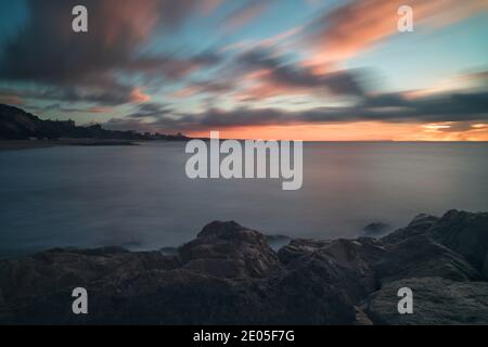 A long exposure captures clouds streaking through the sky as the sun rises into a cold, pale blue winter sky over Bournemouth, Dorset. Stock Photo