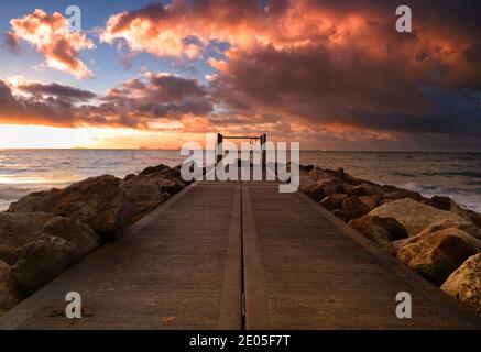 The leading lines of a path flanked by rocks lead out toward the horizon as the rising sun fills a cloudy sky with warm golden and orange colours. Stock Photo