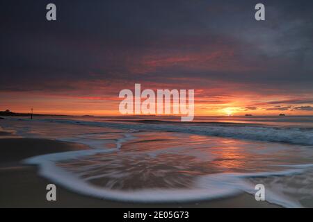 An incoming wave is caught in motion as it advances over the sand of Bournemouth beach under a sky painted orange and red by the rising winter sun. Stock Photo