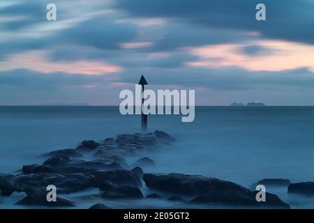 A long exposure time captures the sea washing over the large rocks of a sea defence groyne as the sun rises into a subdued sky on a cold winter day. Stock Photo