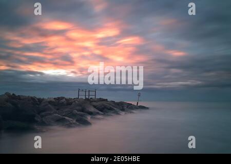 A long exposure time captures the sea washing over the large rocks of a sea defence groyne as the sun rises into a subdued sky on a cold winter day. Stock Photo