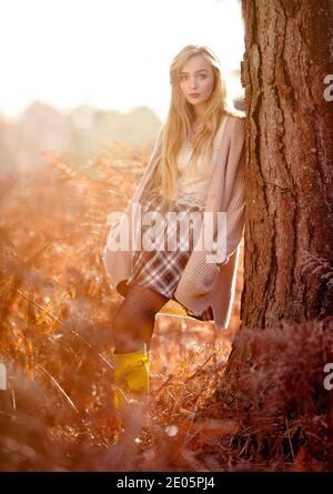 A young beautiful woman (20) dreamy editorial fashion shot in a warm autumnal forest setting wearing a cardigan and tartan skirt with long blonde hair Stock Photo