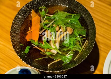 Mizuna leaves, enoki mushrooms and carrot strips simmer in clear broth in a black pot. The vegetables form part of the shabu-shabu hot pot. Restaurant in Shizuoka, Shizuoka Prefecture, Japan. Stock Photo