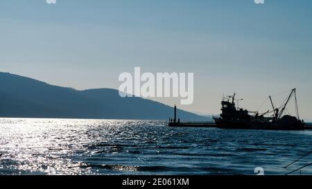 Marmaris, Akyaka, Turkey - November 2020: Silhouette of a fishing ship with unidentified people and a lighthouse in Akyaka village harbor Stock Photo