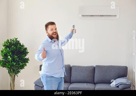 Funny bearded man turning on air conditioner with remote control and showing thumb up in apartment, copy space Stock Photo