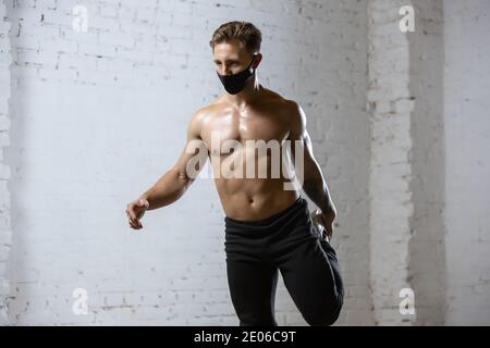 Stretching. Professional male athlete training on brick wall background wearing face mask. Sport during quarantine of coronavirus worldwide pandemic. Young man practicing in gym safe using equipment. Stock Photo