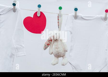 White washed baby clothes, toy and red heart on clothesline with plastic pins. Selective focus, outside view. Stock Photo