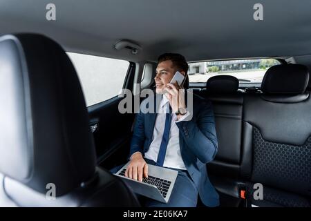 Smiling businessman talking on smartphone and using laptop in car Stock Photo