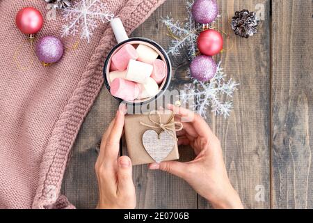 Female hands hold little gift near cup of hot drink with marshmallow on of wooden table with warm knit woolen scarf. Christmas background. Concept of Stock Photo