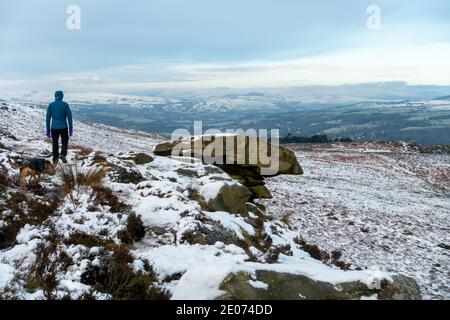 Walker walking a dog next to The Pancake Stone on Ilkley Moor on a snow covered moorland loking towards the Cow and Calf Rocks in West Yorkshire, UK Stock Photo