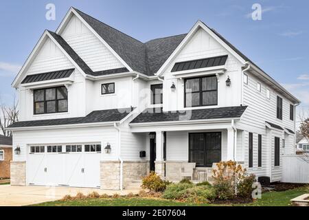 A new, white modern farmhouse with a dark shingled roof and black windows. The bottom of the house has a light rock siding and a covered front porch. Stock Photo