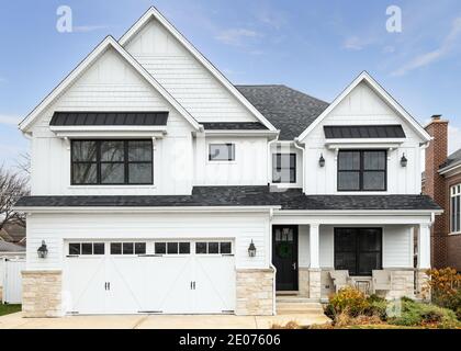 A new, white modern farmhouse with a dark shingled roof and black windows. The bottom of the house has a light rock siding and a covered front porch. Stock Photo