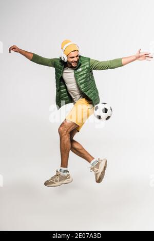 Cheerful sportsman in headphones jumping near football on grey background Stock Photo