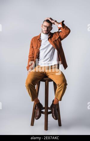 Fashionable man in turtleneck and terracotta jacket looking at camera on wooden chair on grey background Stock Photo