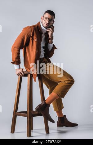 Fashionable man in terracotta jacket and brown shoes posing near wooden chair on grey background Stock Photo
