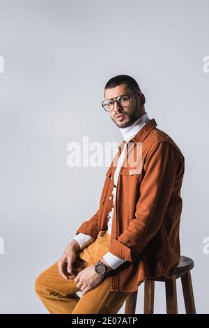 Stylish man in eyeglasses and terracotta jacket sitting on chair isolated on grey Stock Photo