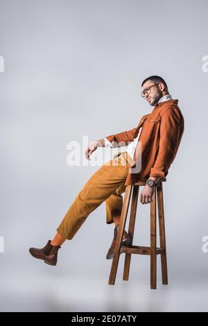 Stylish man in eyeglasses and terracotta jacket posing on wooden chair on grey background Stock Photo