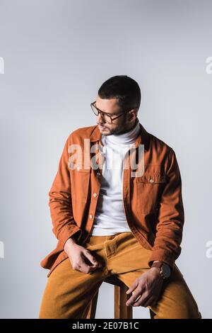 Fashionable man in jacket and eyeglasses sitting on chair and looking away on grey background Stock Photo