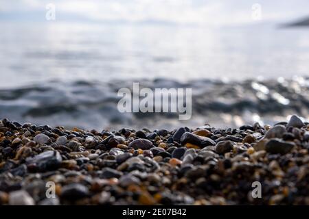 Focus on the middle of many shiny wet and colorful pebbles next to the shore. In background waves and horizon. Sky reflects in water Stock Photo
