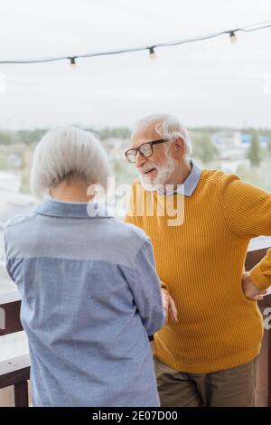 back view of elderly wife standing near smiling husband with hand on hip on terrace on blurred background Stock Photo