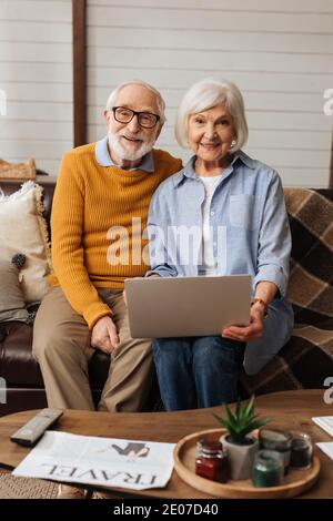 happy elderly couple with laptop looking at camera on couch on blurred foreground in living room Stock Photo