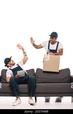 tattooed worker sitting on sofa, holding digital tablet and giving high five to indian mover with box on white Stock Photo