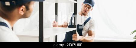 young mover carrying heavy rack with indian coworker on blurred foreground, banner Stock Photo