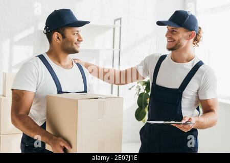 cheerful multicultural movers smiling while looking at each other in apartment Stock Photo