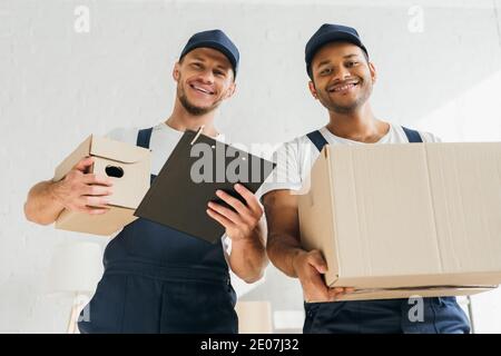 low angle view of cheerful multicultural movers smiling while looking at camera and holding boxes in apartment Stock Photo