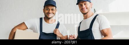 cheerful multicultural movers smiling while looking at camera in apartment, banner Stock Photo