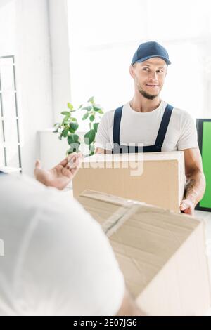 surprised worker in cap and uniform carrying box while looking at indian coworker on blurred foreground Stock Photo