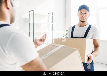 surprised mover in cap and uniform carrying box while looking at indian coworker on blurred foreground Stock Photo