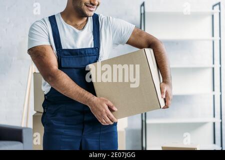 cropped view of cheerful indian courier in uniform holding box in apartment Stock Photo