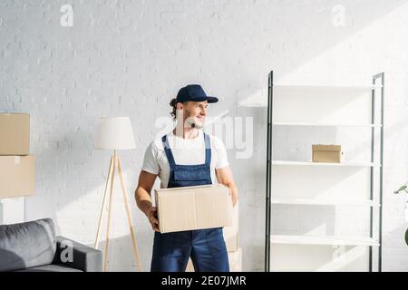 happy mover in cap holding carton box and looking away in apartment Stock Photo