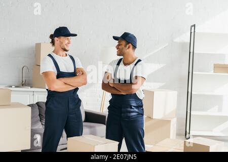 cheerful movers in caps standing with crossed arms while looking at each other near boxes in room Stock Photo