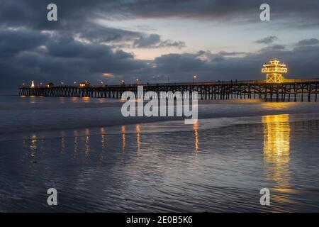 A brightly lit ocean pier on a cold December evening casts reflections on the smooth beach waters below. Stock Photo