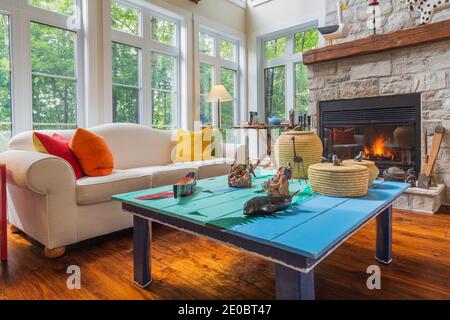 White upholstered sofa with colourful velour cushions, folk art coffee table, grey and beige cut stone wood burning fireplace in family room Stock Photo