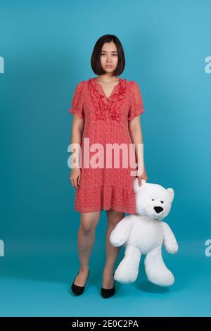 full-length portrait of a sad, upset, offended, lonely woman in a red dress holding a white Teddy bear by the ear, isolated on a blue background